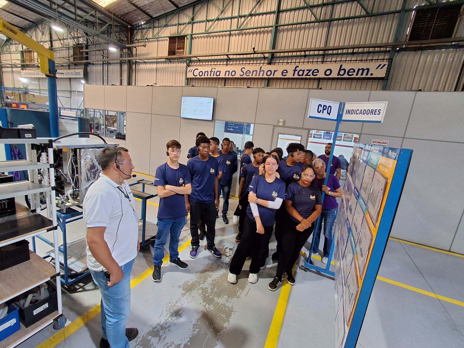 Um grupo de pessoas ouve um homem em uma fábrica, enquanto ele observa um painel de exibição.