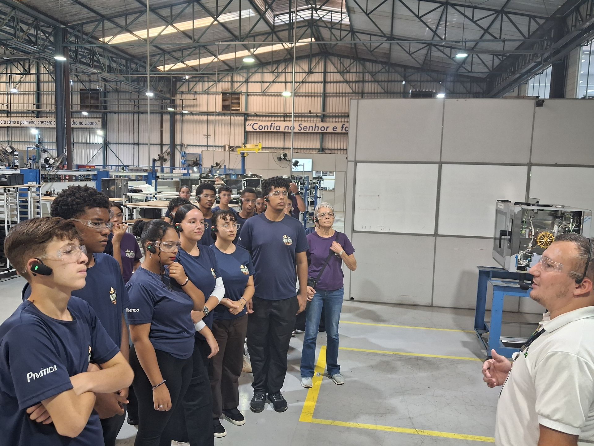Grupo de pessoas em um ambiente fabril, ouvindo um homem. A maioria veste camisas azuis.