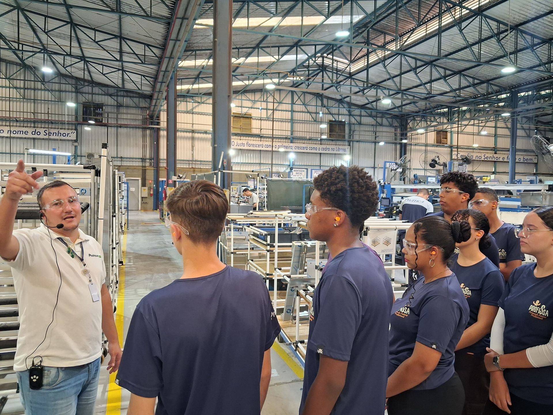 O guia turístico gesticula enquanto explica o funcionamento dos equipamentos da fábrica a um grupo de estudantes.