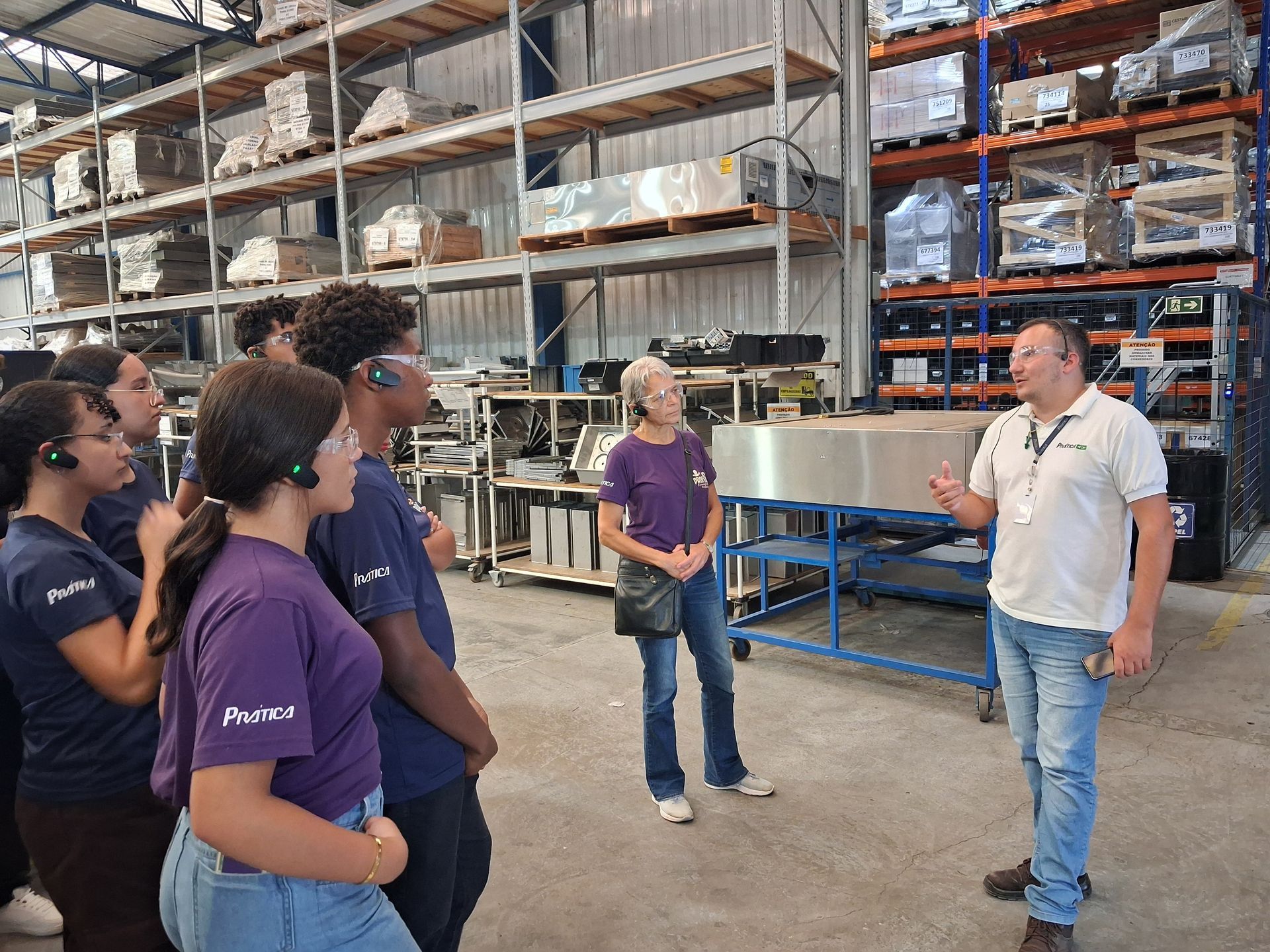 Visita guiada em grupo a um armazém: um homem fala, apontando para os equipamentos, enquanto outros ouvem atentamente, usando fones de ouvido. Prateleiras repletas de itens.