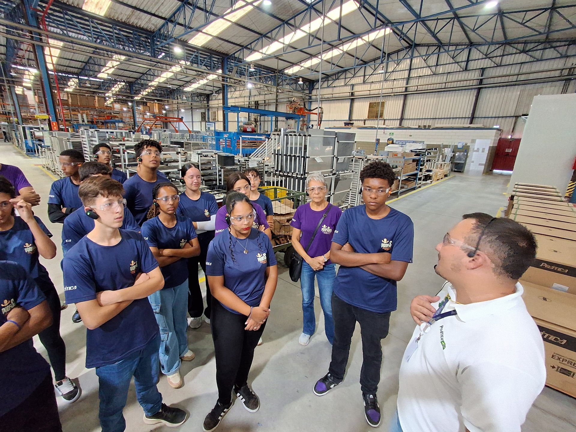 Um grupo de estudantes com camisas azuis ouve um palestrante em um ambiente fabril.