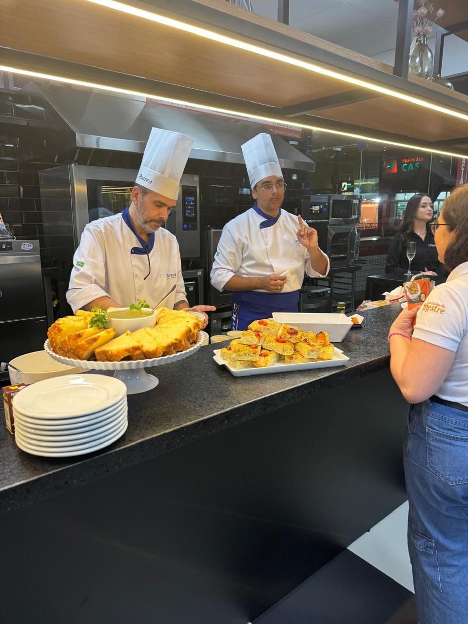 Dois chefs estão preparando comida em uma cozinha enquanto uma mulher observa.