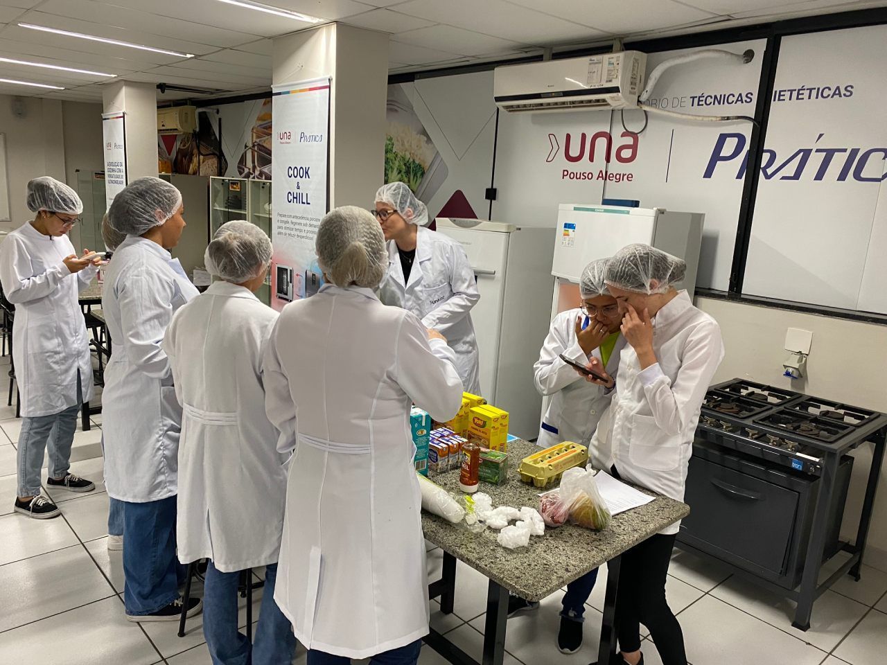 Um grupo de pessoas com jalecos e redes de cabelo estão em pé ao redor de uma mesa.