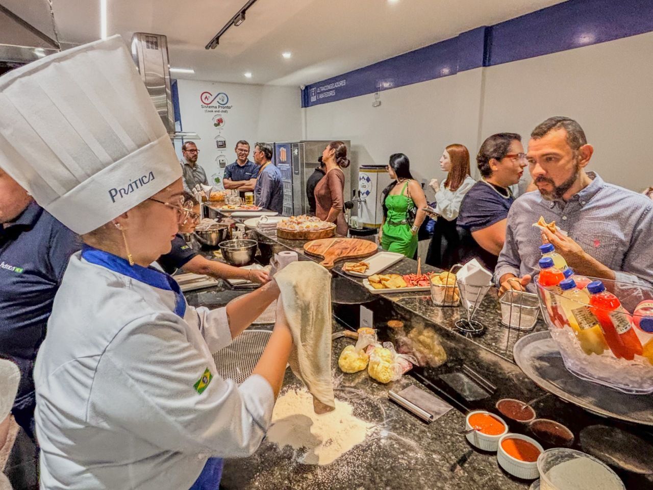 Chef de chapéu alto esticando massa em um balcão com uma multidão de pessoas em uma cozinha bem iluminada.