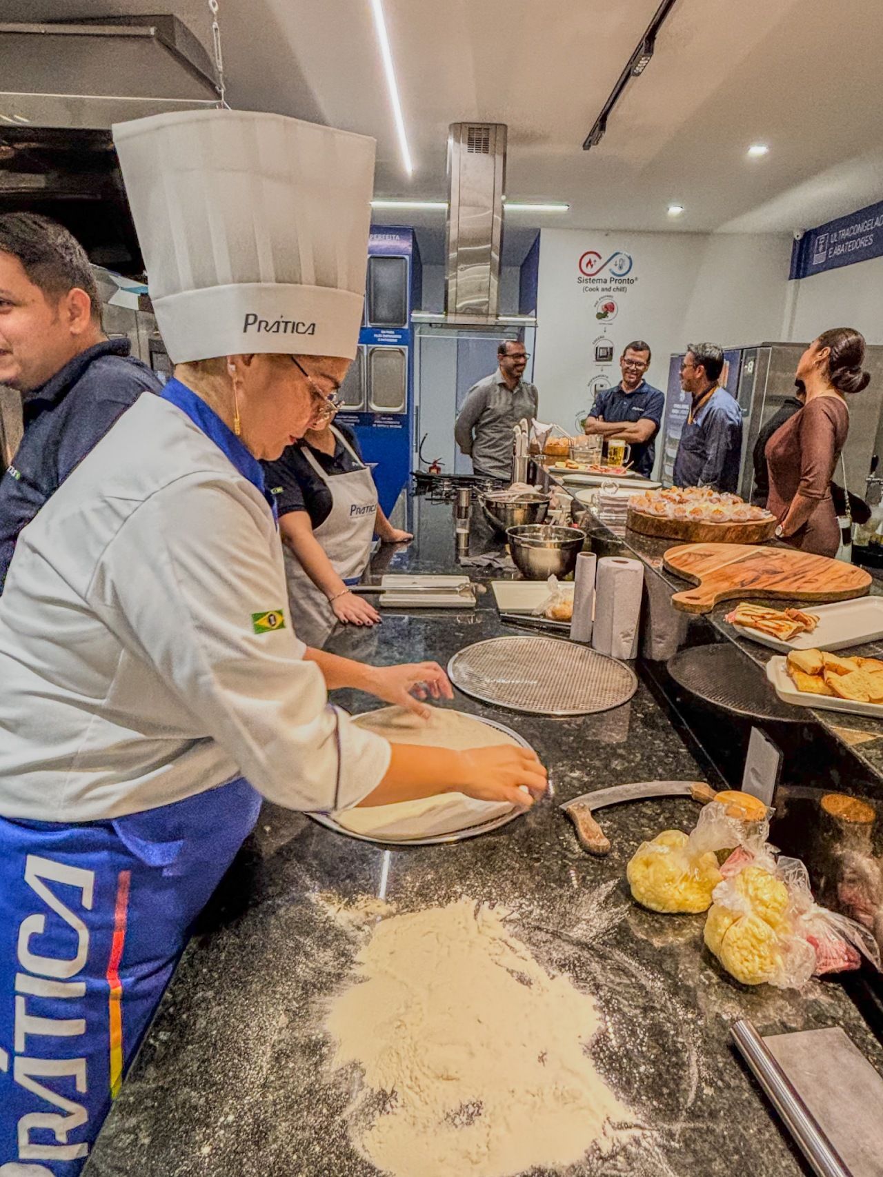 Chef de cozinha com chapéu e avental brancos preparando massa em uma bancada, com outras pessoas ao fundo.