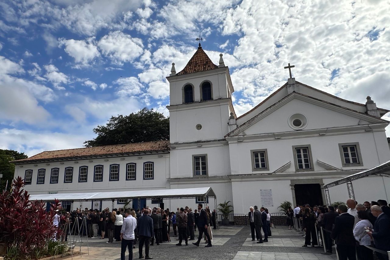 Igreja branca com uma cruz, pessoas reunidas do lado de fora em um dia ensolarado.