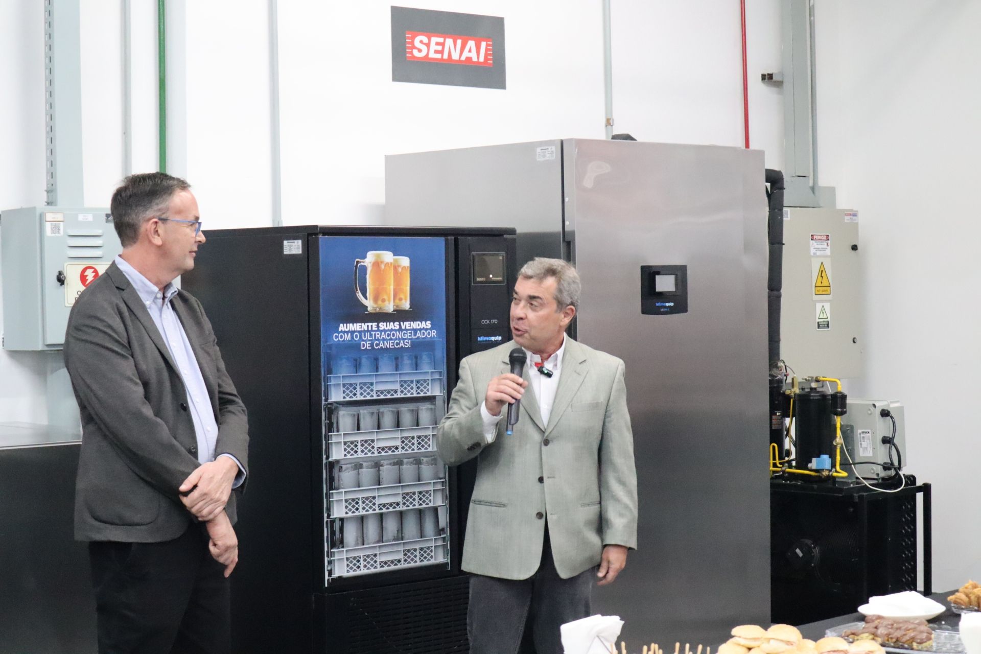 Dois homens em uma sala; um fala em um microfone perto de uma máquina de venda automática, enquanto o outro observa. Uma placa da SENAI é visível.