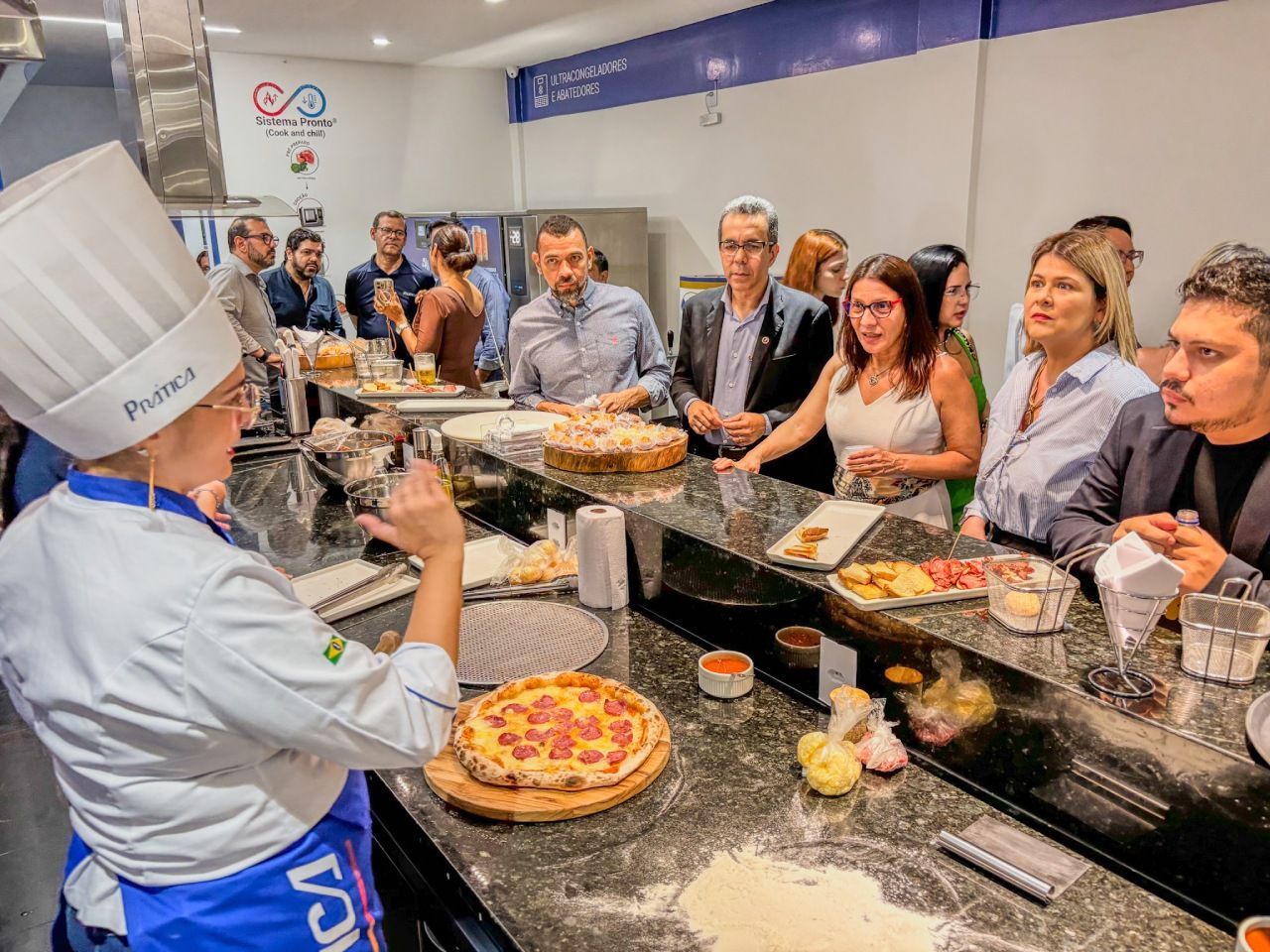 O chef demonstra como fazer pizza para um grupo em uma cozinha. A pizza, os ingredientes e os utensílios estão sobre a bancada.