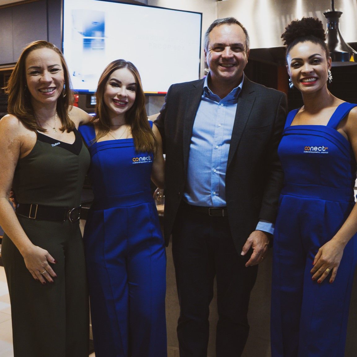 Um homem de terno e duas mulheres de macacão azul estão posando para uma foto