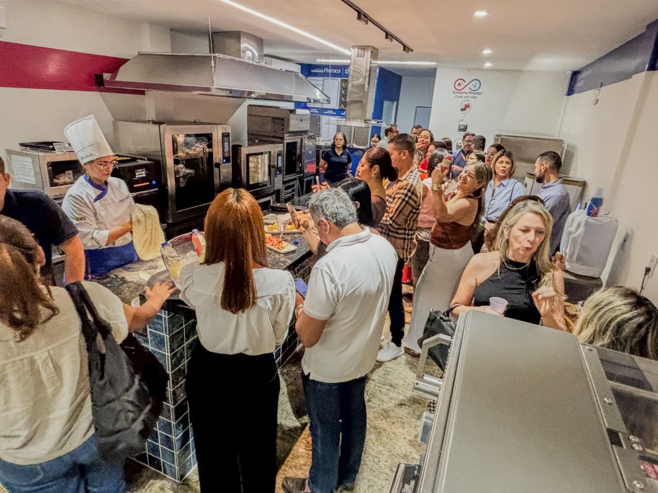 Chef preparando comida em uma cozinha, cercado por um grupo de pessoas observando. Cozinha comercial bem iluminada.