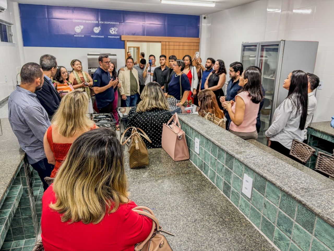 Um grupo de pessoas estava reunido em uma sala, algumas ouvindo um orador, perto de uma bancada e de uma porta.