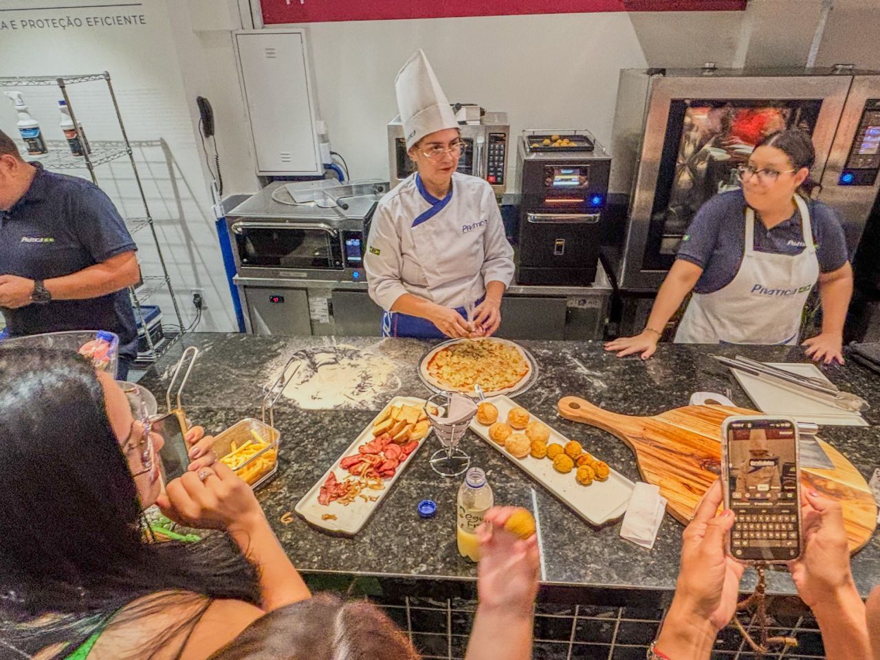 Um chef de chapéu branco prepara comida em um balcão, com assistentes e pessoas tirando fotos.