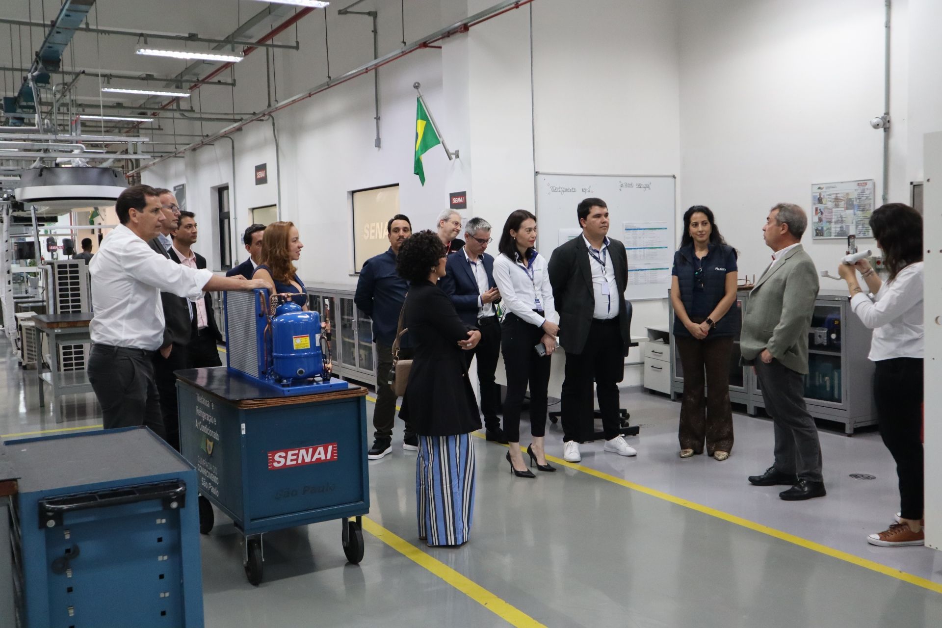 Grupo de pessoas visitando uma fábrica. Elas estão assistindo a uma apresentação perto de máquinas azuis.