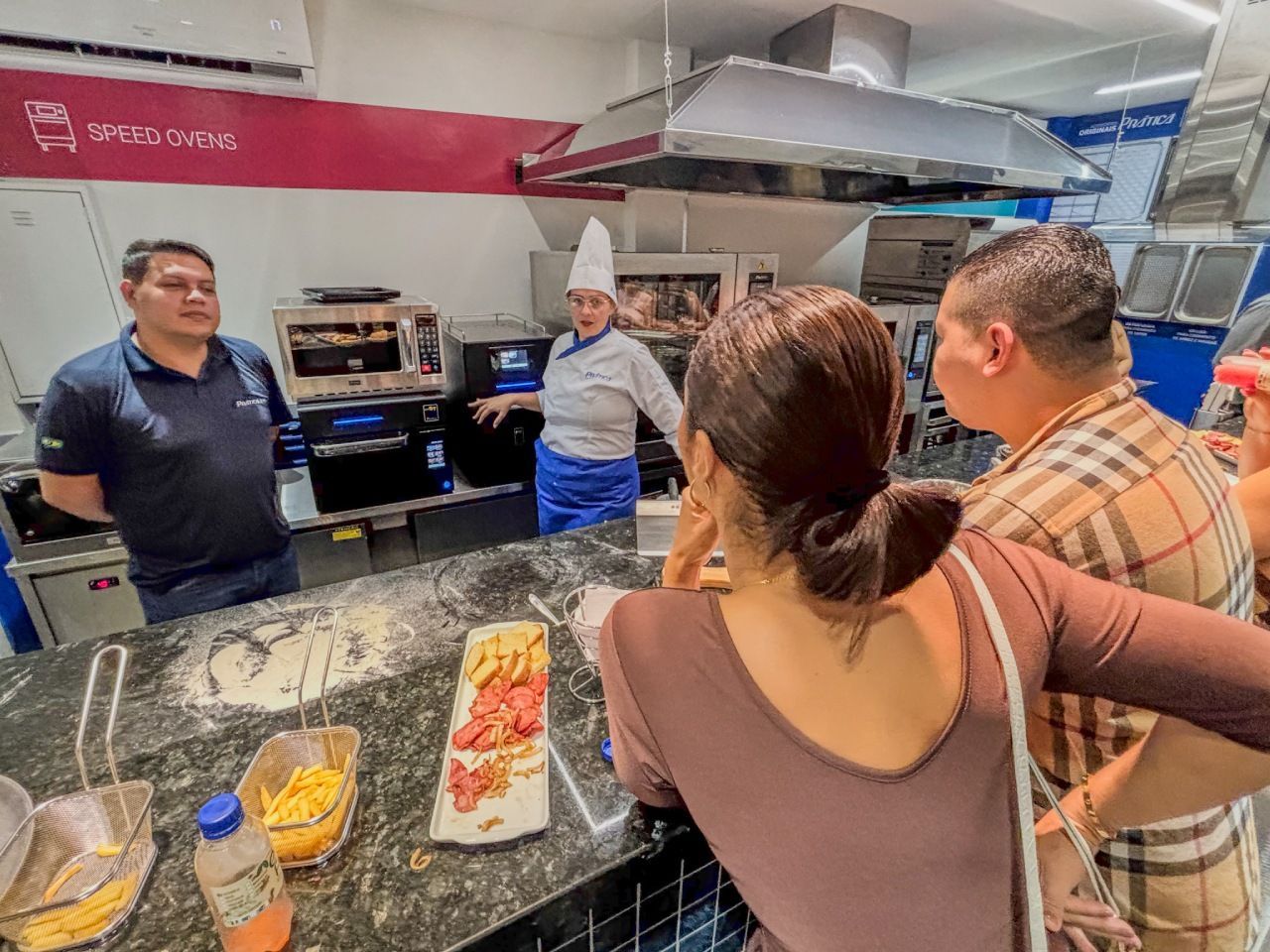 Pessoas assistem a um chef demonstrando o uso de eletrodomésticos em um showroom. Um balcão exibe amostras de alimentos.