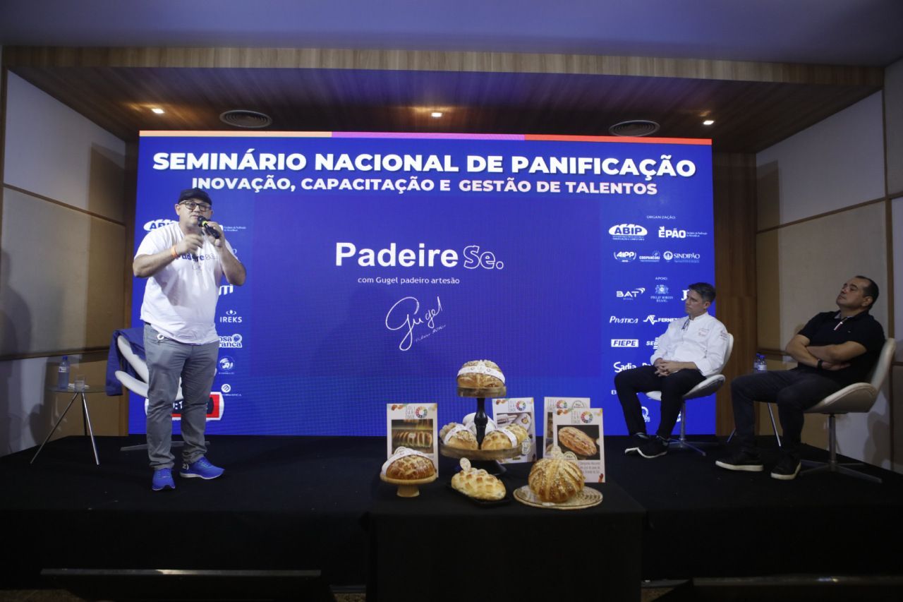 Dois homens estão sentados em frente a uma tela que diz seminário nacional de panificação