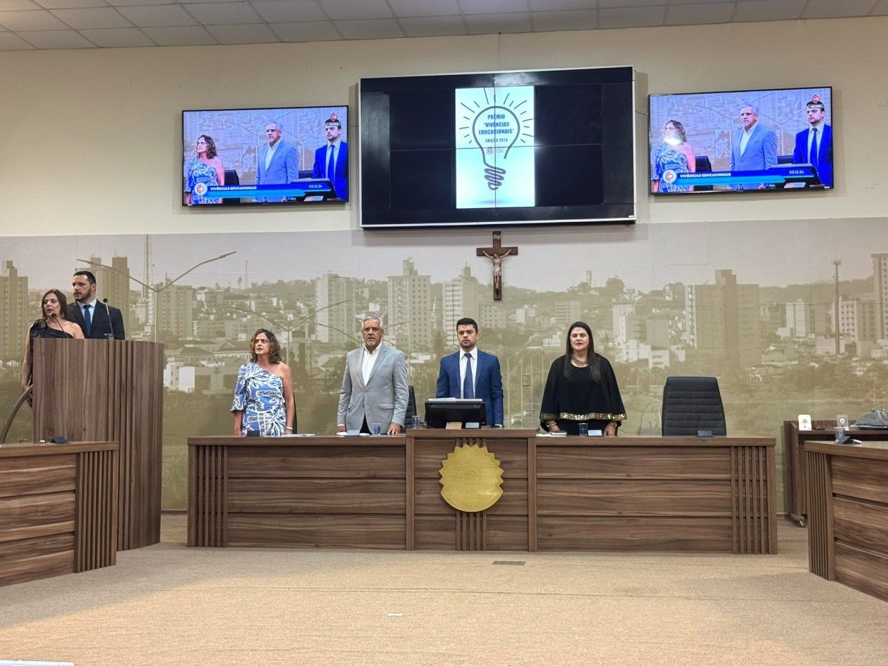 Um grupo de pessoas em pé em frente a um pódio em uma câmara do conselho.
