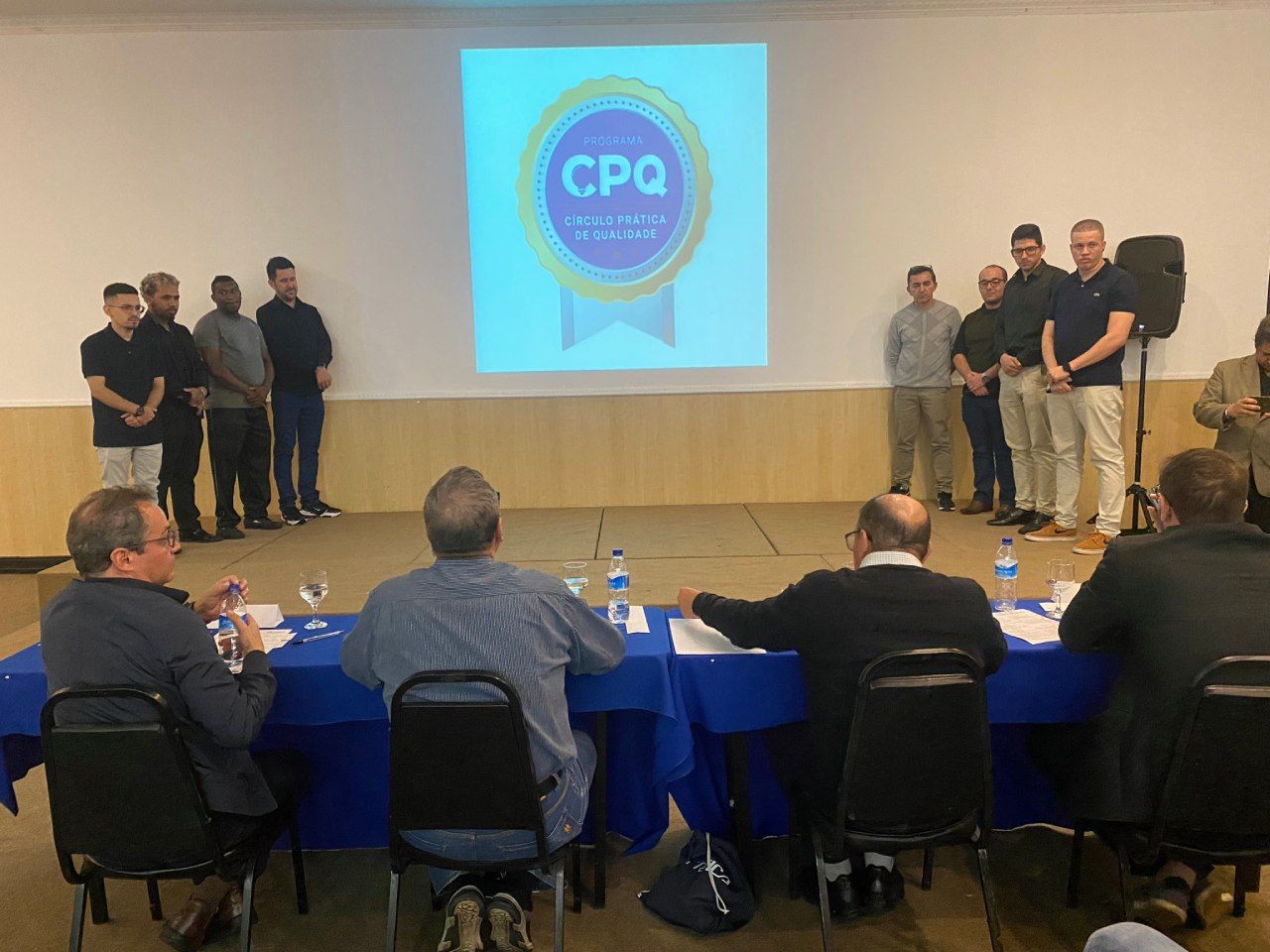 Um grupo de homens está sentado em mesas em frente a uma tela que diz cpq.