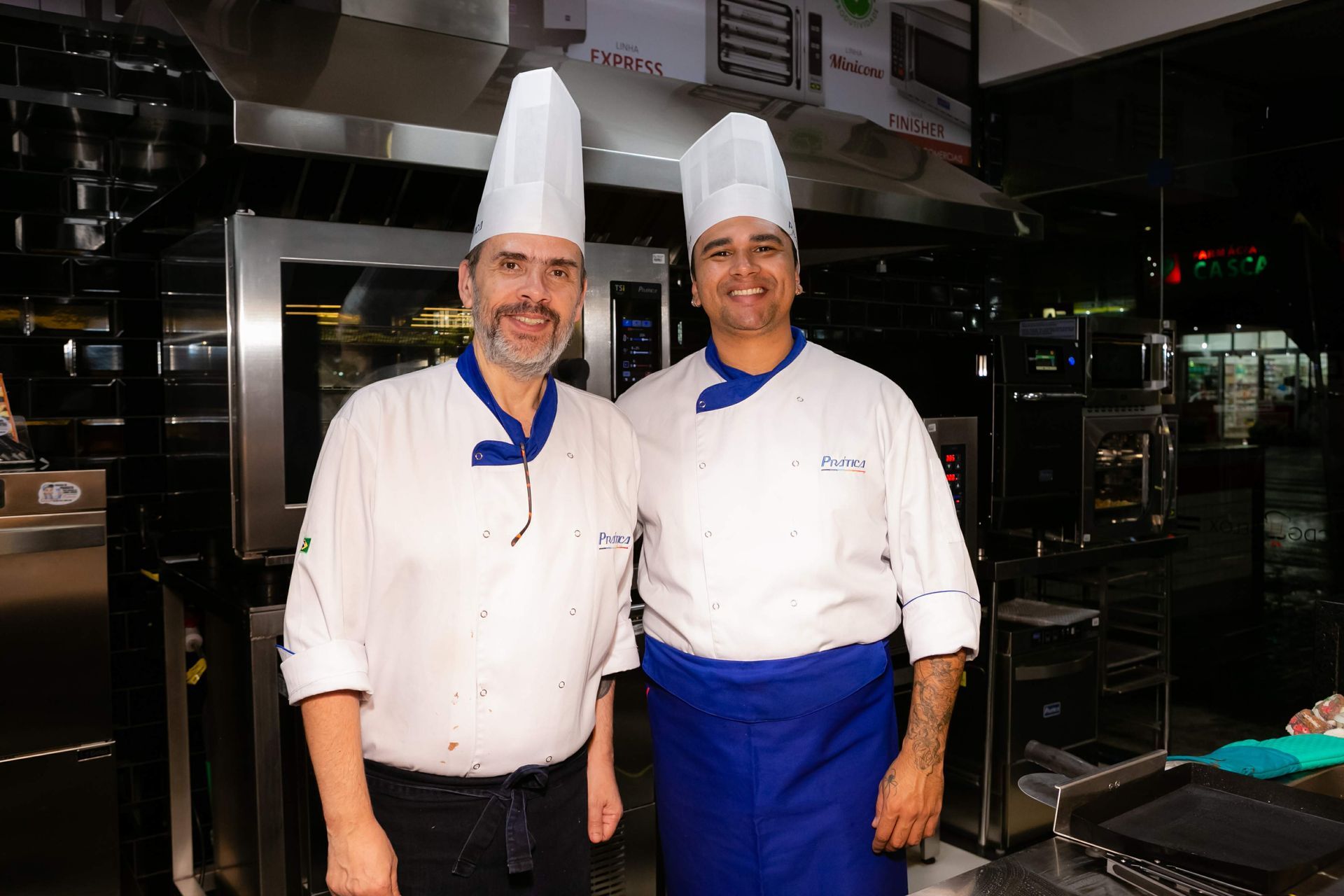 Dois chefs estão de pé, um ao lado do outro, em uma cozinha.