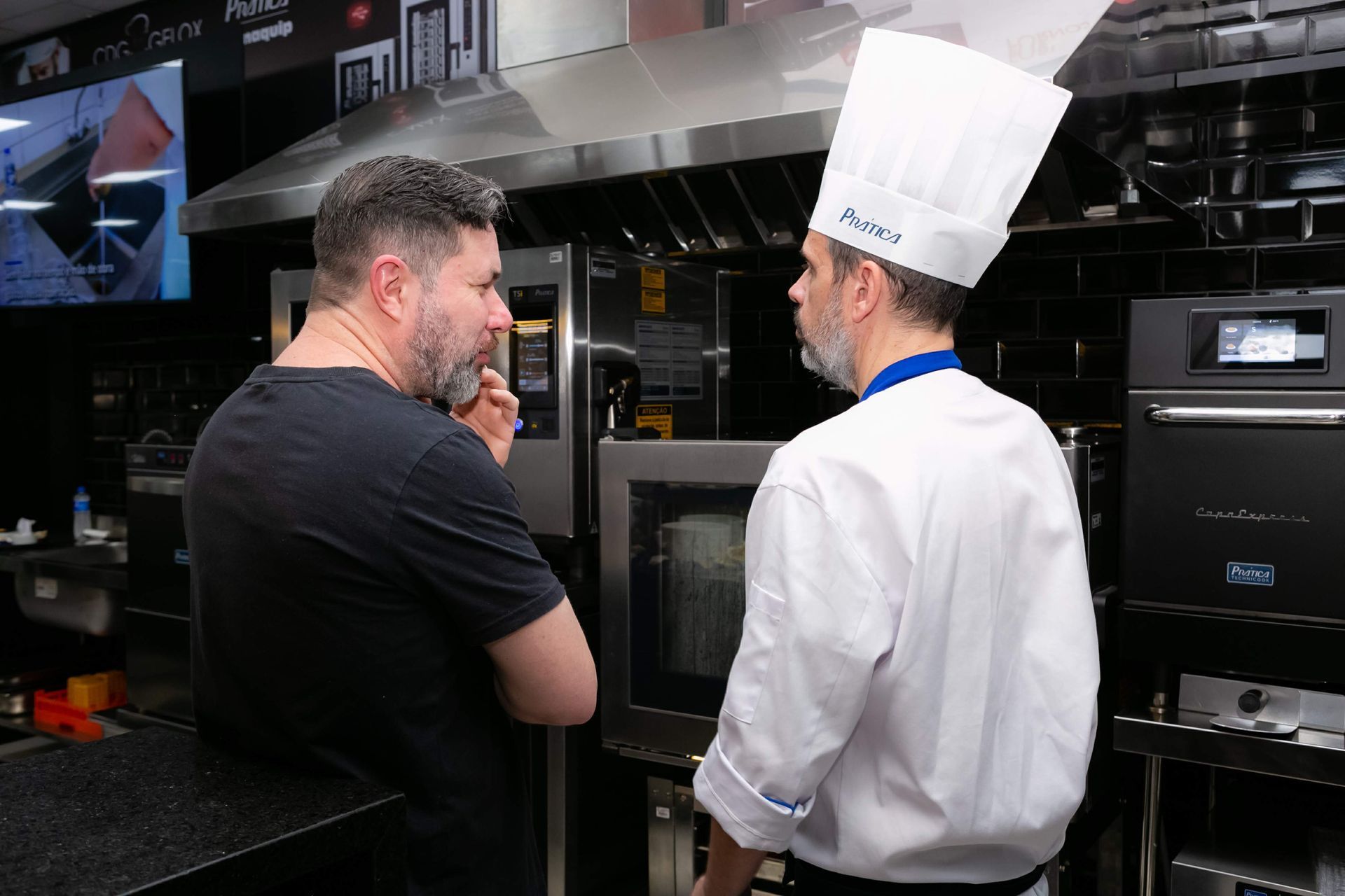 Dois homens estão parados um ao lado do outro em uma cozinha conversando entre si.