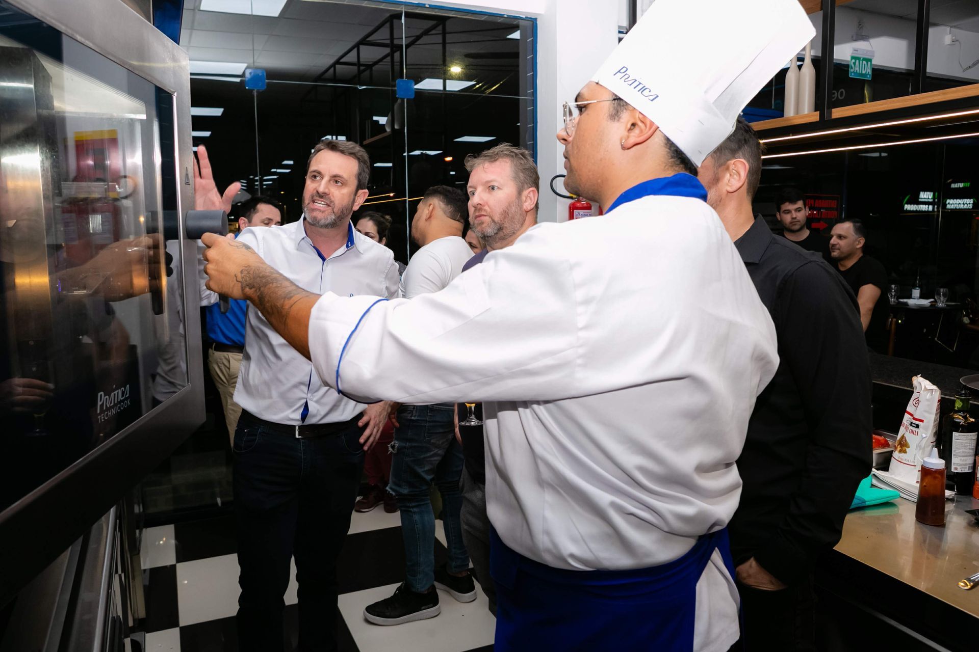Um grupo de pessoas está em volta de um chef em uma cozinha.