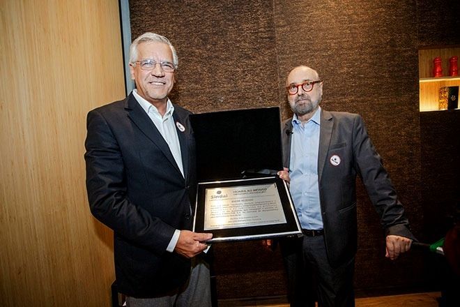 Dois homens seguram uma placa emoldurada em um ambiente interno. O homem à esquerda sorri enquanto o outro observa a placa.