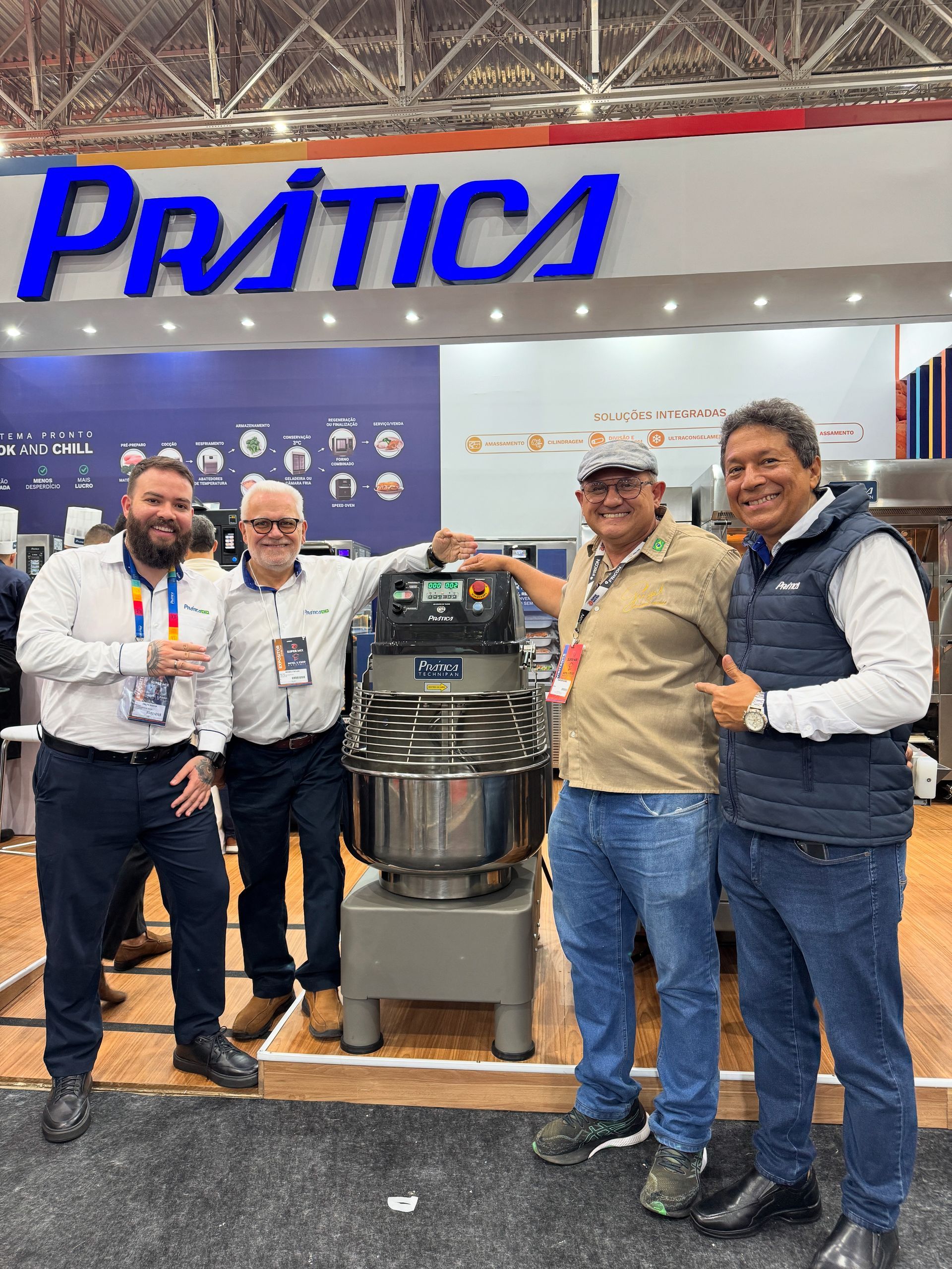 Quatro homens estão ao lado de um grande misturador industrial em um estande de feira comercial, com a placa 
