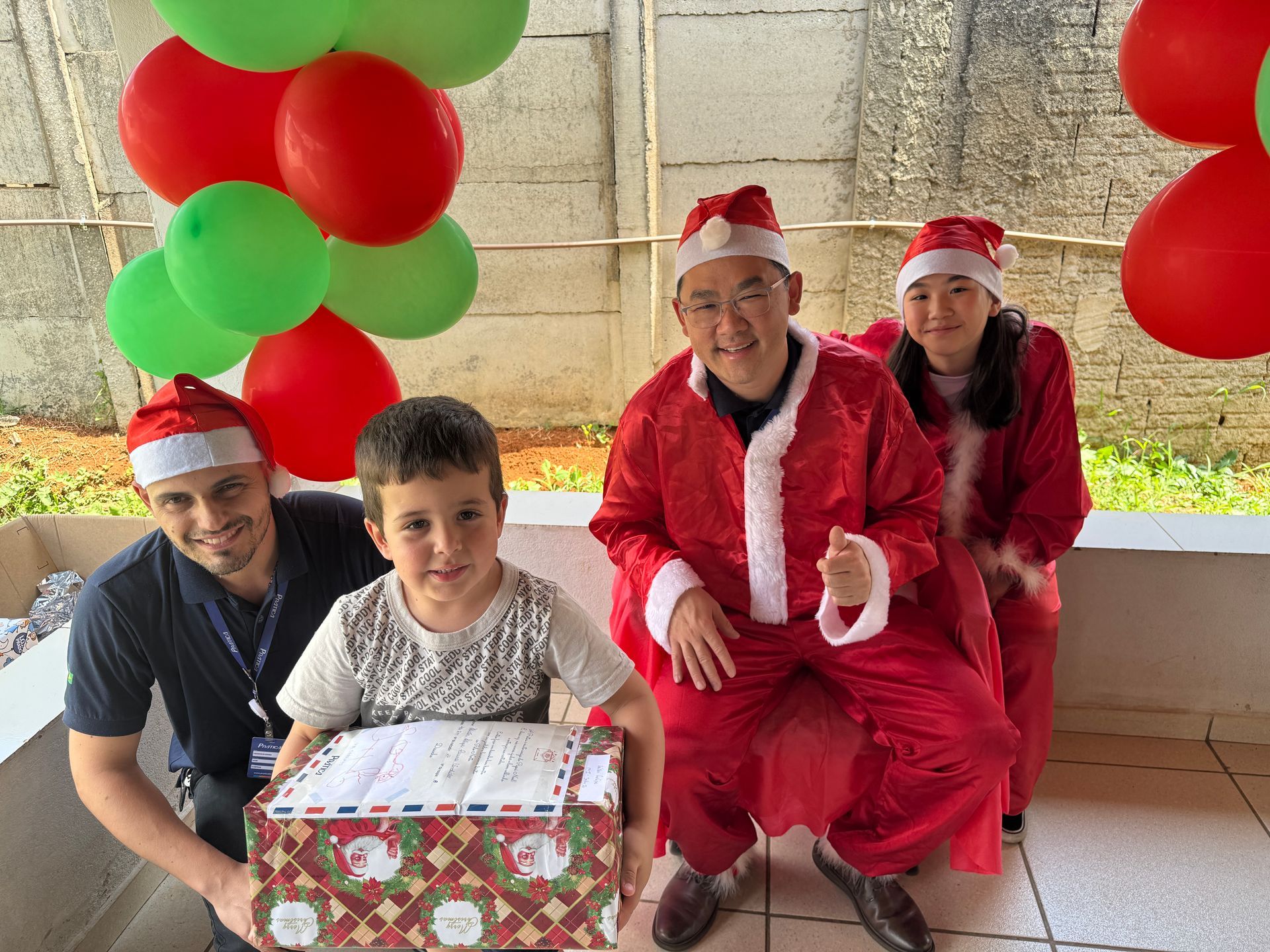Pessoas com gorros e trajes de Papai Noel, segurando balões e um presente de Natal, posando ao ar livre.