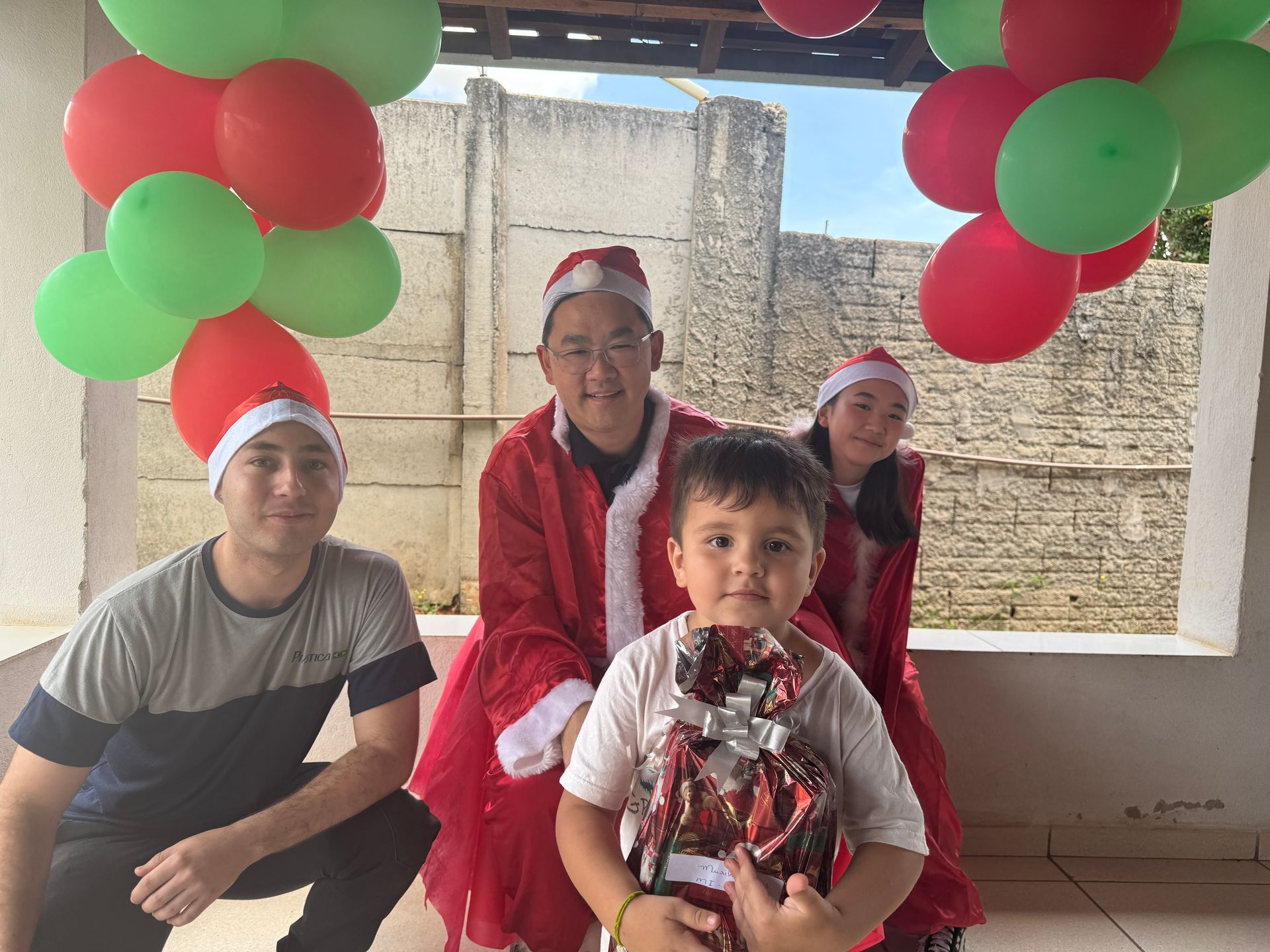 Pessoas com gorros de Papai Noel posam com balões, e uma criança segura um presente. Cenário festivo ao ar livre.