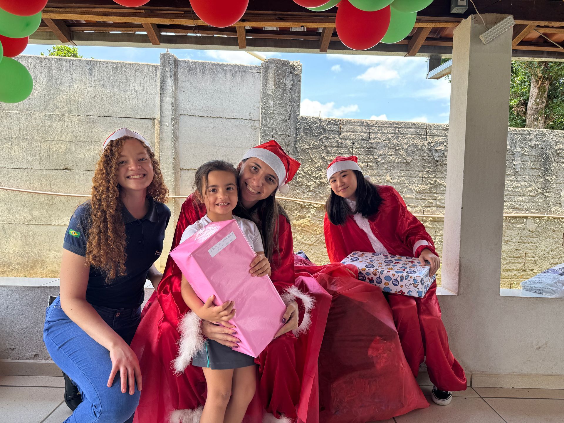 Pessoas com gorros de Papai Noel entregam presentes a uma criança. Balões vermelhos e verdes decoram uma varanda.