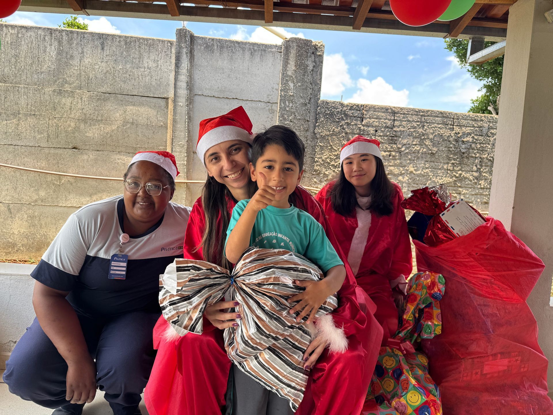 Quatro pessoas com gorros de Papai Noel posam com uma criança segurando um grande laço, com presentes por perto.