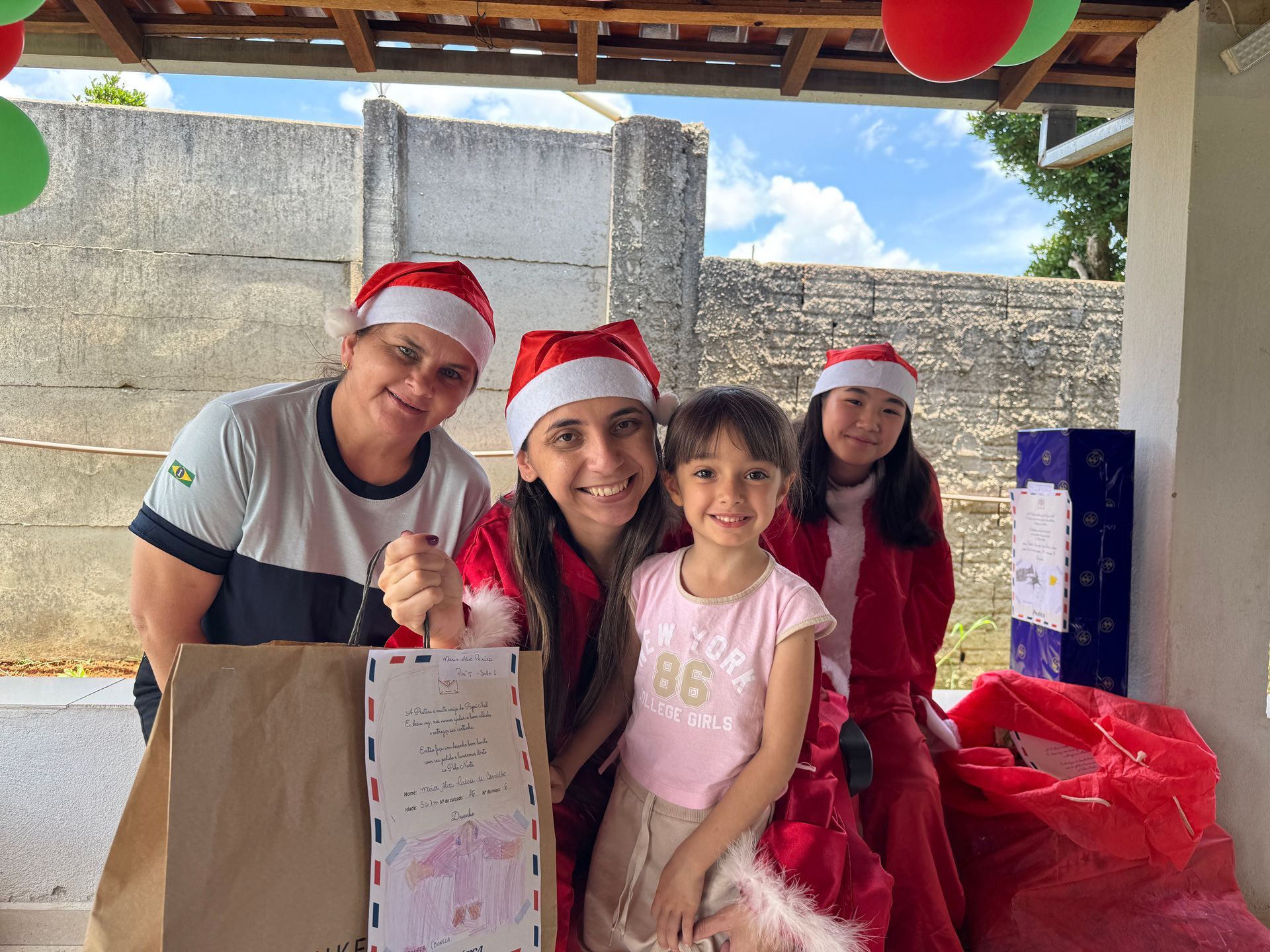 Quatro pessoas com gorros de Papai Noel posam com presentes do lado de fora.