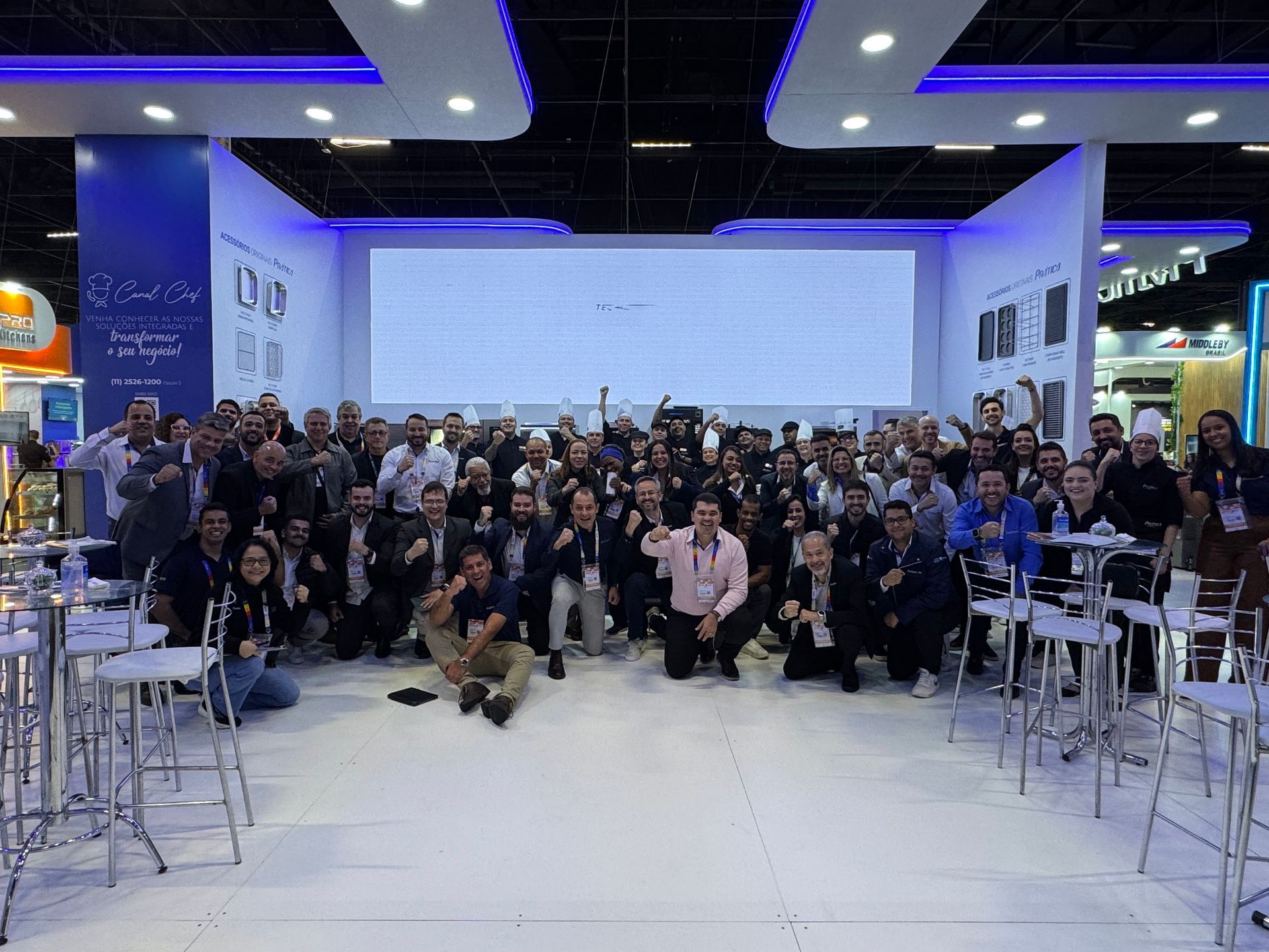 Um grande grupo de pessoas está posando para uma foto em uma sala com mesas e cadeiras.