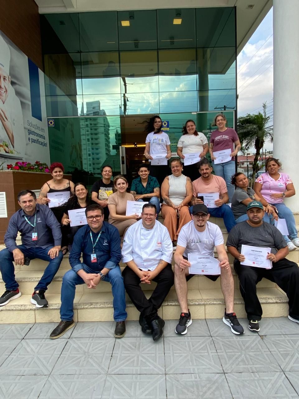 Grupo de pessoas segurando certificados, posando em uma escadaria em frente a um prédio com portas de vidro.