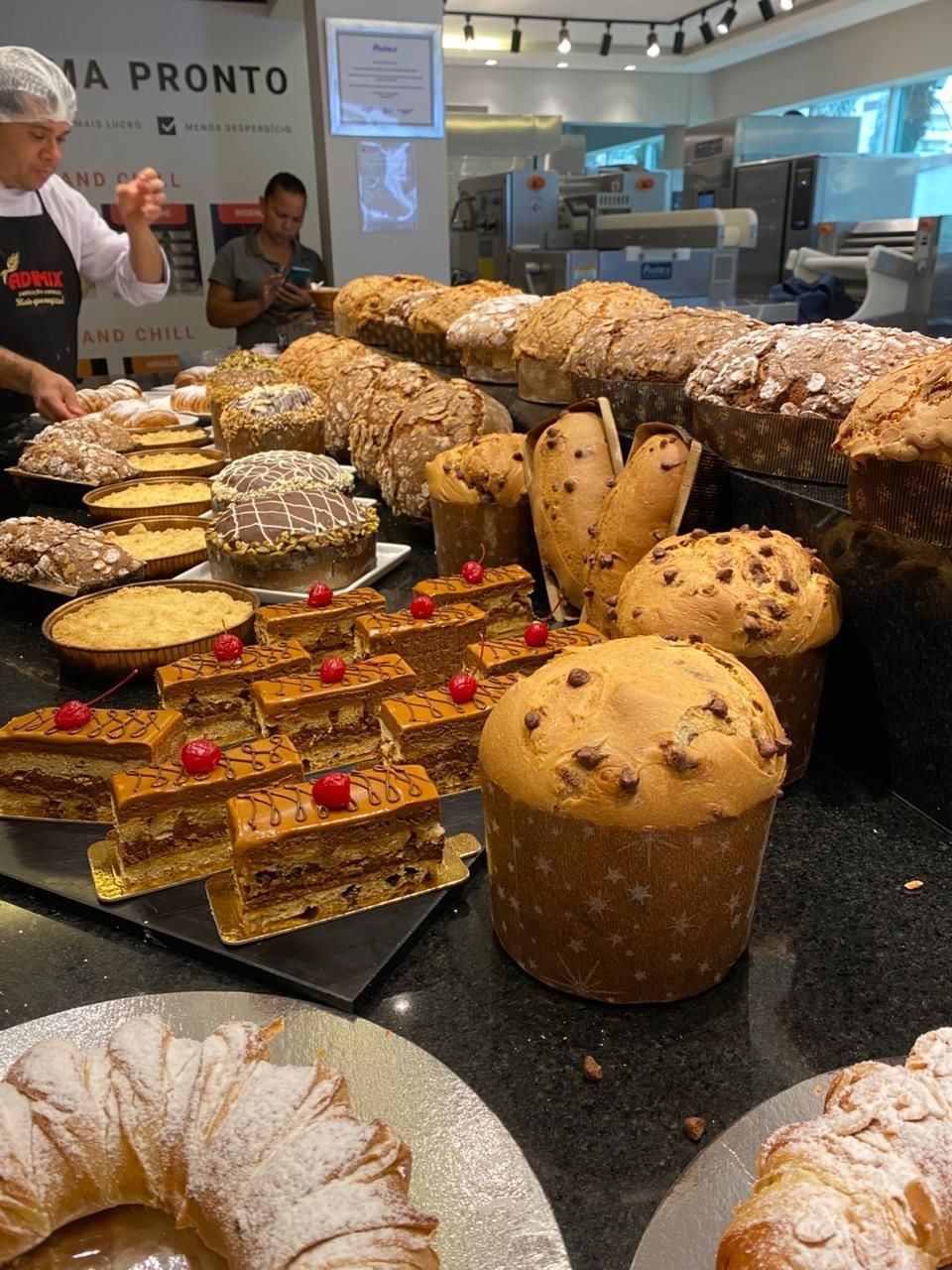 Vitrine de padaria com diversos produtos assados: bolos, pães, doces. Um chef está atrás do balcão.