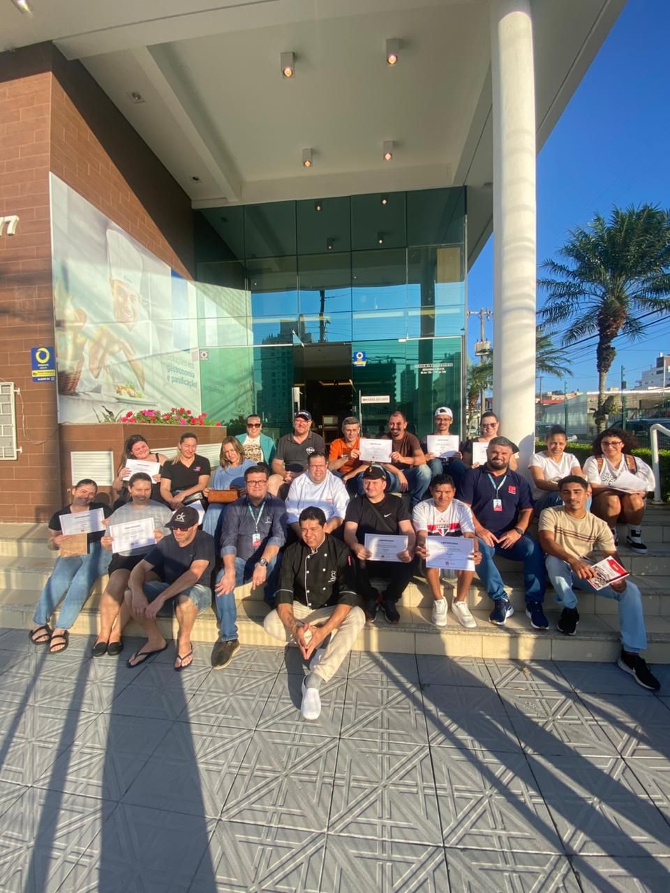 Grupo de pessoas posando na escadaria de um prédio com portas de vidro e uma coluna. Dia ensolarado.