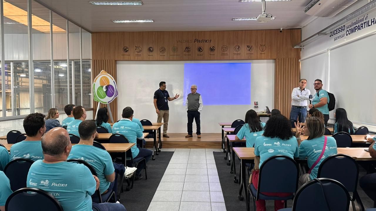 Pessoas em uma sala de aula, com palestrantes em um pequeno palco. Os participantes estão sentados e vestindo camisas turquesa.