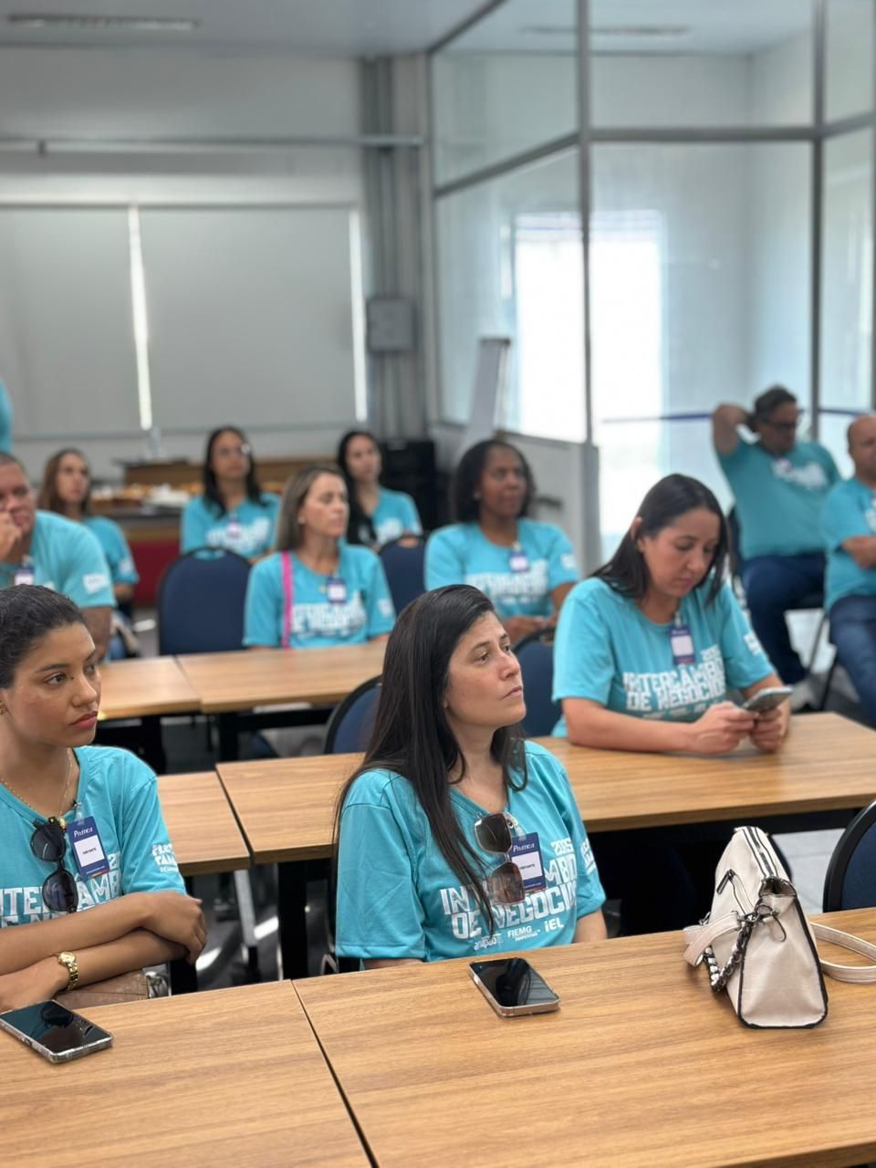 Pessoas vestindo camisetas verde-azuladas sentadas em uma sala de aula, ouvindo atentamente uma apresentação.