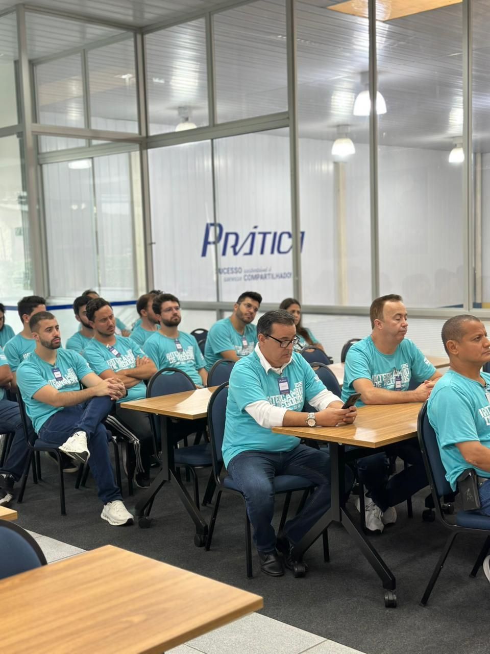 Grupo de homens com camisas verde-azuladas iguais, sentados em carteiras numa sala de aula, ouvindo. Logotipo 