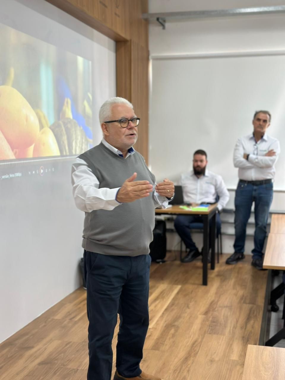 Homem falando e gesticulando em uma sala com outros dois homens. Uma tela de projeção está atrás dele.