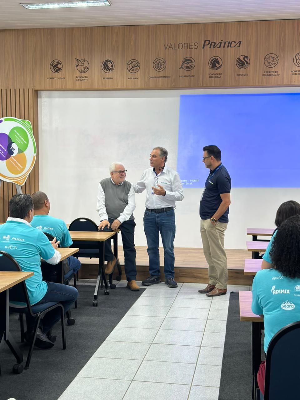 Três homens estão em um pequeno palco falando para uma plateia sentada em uma sala de aula.