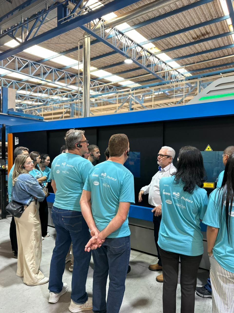 Um grupo de pessoas vestindo camisetas verde-azuladas iguais assiste a uma apresentação perto de equipamentos industriais.