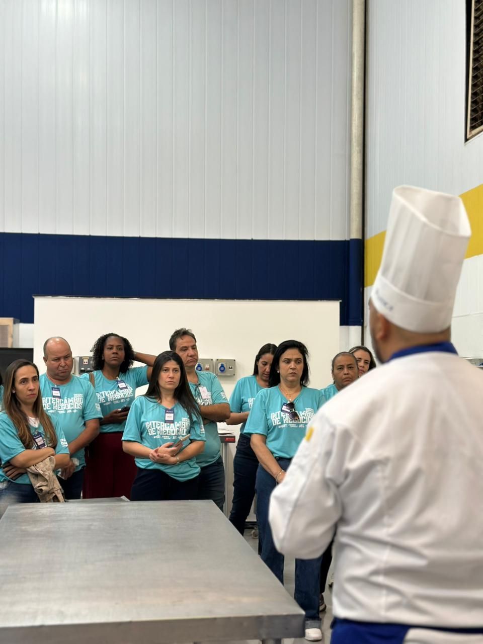 Chef falando com um grupo de pessoas vestindo camisetas verde-azuladas dentro de uma cozinha industrial.