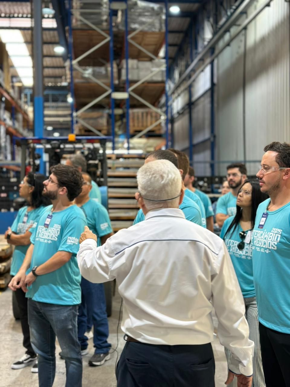 Grupo de pessoas em um armazém, ouvindo um homem de jaqueta branca e camisa azul, perto de prateleiras altas.