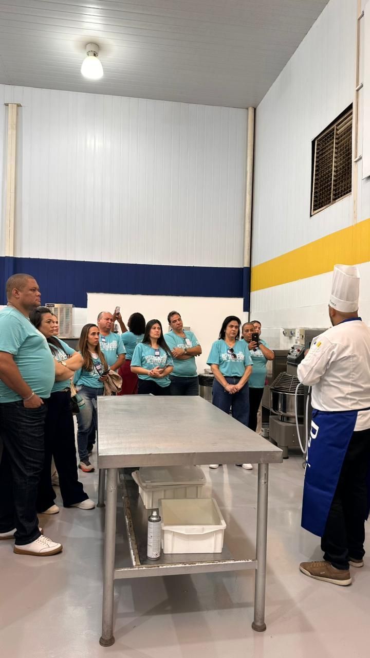 Um chef fazendo uma apresentação para um grupo em uma cozinha. O grupo veste camisetas verde-azuladas e está de frente para o chef.