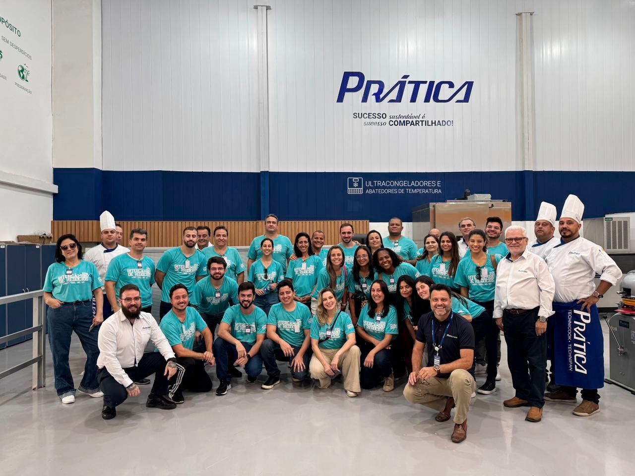 Grupo de pessoas vestindo camisetas verde-azuladas posando em frente ao logotipo da Prática, dentro de uma fábrica.