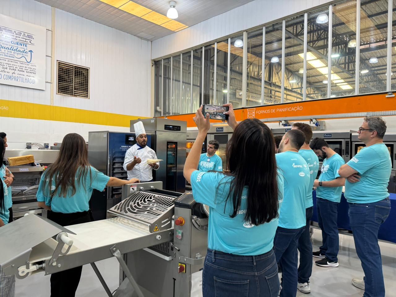 Grupo de pessoas assistindo a uma demonstração culinária em uma padaria. Algumas estão tirando fotos com seus celulares.