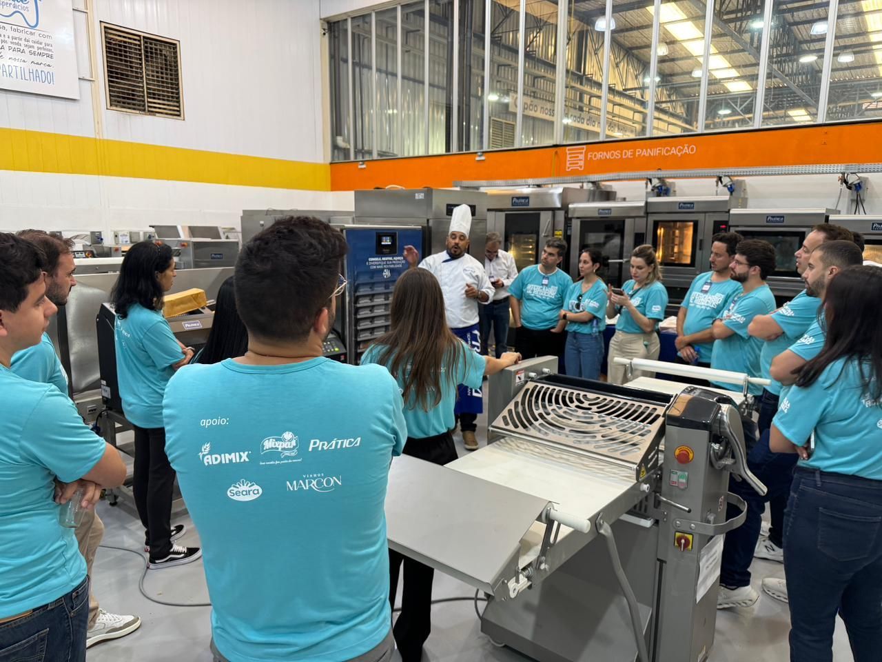 Um grupo de pessoas vestindo camisas azuis observa um chef em uma cozinha, perto de equipamentos industriais de panificação.