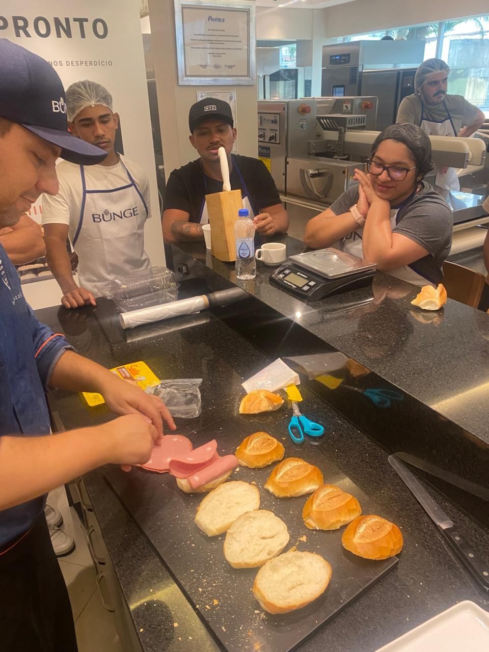 Padeiro preparando sanduíches em uma cozinha, com várias pessoas observando.