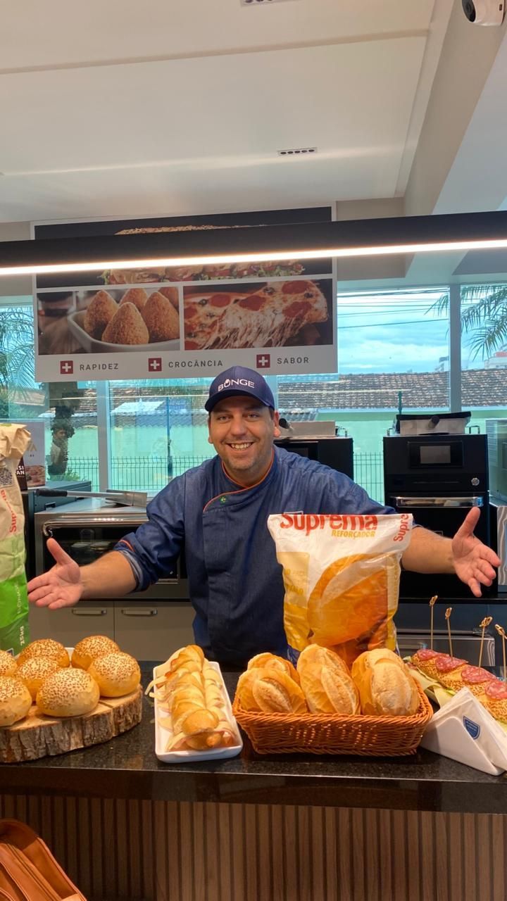 Homem de avental azul de chef em um balcão de padaria, braços estendidos, sorrindo. Pães e doces expostos.