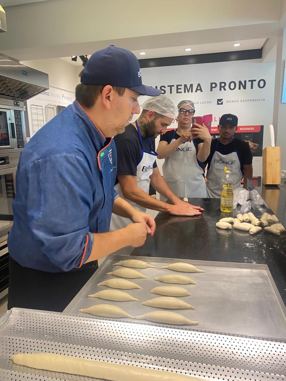 Chef demonstra como moldar pães para um grupo em uma cozinha. Os pãezinhos estão em uma bandeja e em uma bancada.